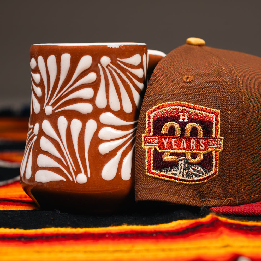 A brown mug with white floral patterns sits next to a Bendicion de la Abuela 20th Brown Red Houston Astros New Era Hat 5950 Fitted, both resting on a colorful striped cloth.