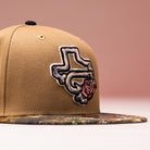 The La Ofrenda Khaki RTC Copa Corpus Christi Hooks New Era 5950 Fitted hat features an embroidered Texas outline and a red rose on the front, with a camouflage-patterned brim, shown against a soft pink background.