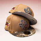 Two La Ofrenda Khaki RTC Copa Corpus Christi Hooks New Era 5950 Fitted hats, featuring Texas-shaped logos with roses, side patches, and camo brims, stacked atop each other on a pink gradient background. Perfect for fans!.