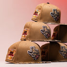 Five La Ofrenda Khaki RTC Copa Corpus Christi Hooks New Era 5950 fitted hats are stacked in two tiers. Each features multicolored embroidered Texas and trophy patches, set against a pink gradient background.