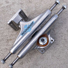 Two Legacy OGBC INDY 139 Polished Silver Standard Stage 11 skateboard trucks are stacked in an X shape on a rough, light-colored concrete surface. The kingpin and bushings are visible on the lower truck, highlighting the durable aluminum construction of these standard Stage 11 trucks built to handle every ride.