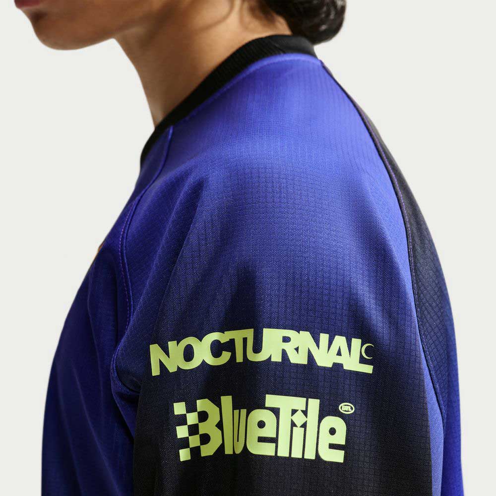 A person wears the Nike SB Skateboarding Long Sleeve Skate Top IQ6594514, a blue and black jacket with “NOCTURNAL” and “BlueTile” in light yellow on the upper sleeve, featuring an imported polyester rib finish, shown from the side.