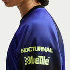 A person wears the Nike SB Skateboarding Long Sleeve Skate Top IQ6594514, a blue and black jacket with “NOCTURNAL” and “BlueTile” in light yellow on the upper sleeve, featuring an imported polyester rib finish, shown from the side.