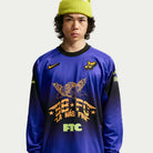 A young person with curly hair wears the Nike SB Skateboarding Long Sleeve Skate Top IQ6594514, featuring blue, black, and gold graphics including SB FC LA MÁS FINA and FTC. The imported polyester top stands out against a plain light background.