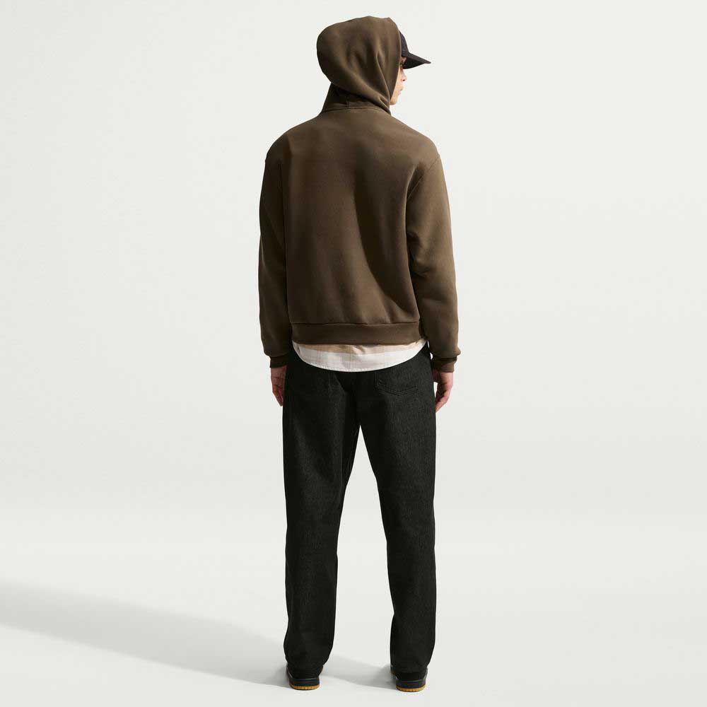 A person faces away wearing the Nike SB Loose Denim Black Pant IB2992010, brown hoodie with hood up, black cap, white shirt showing, and brown shoes against a plain white background.