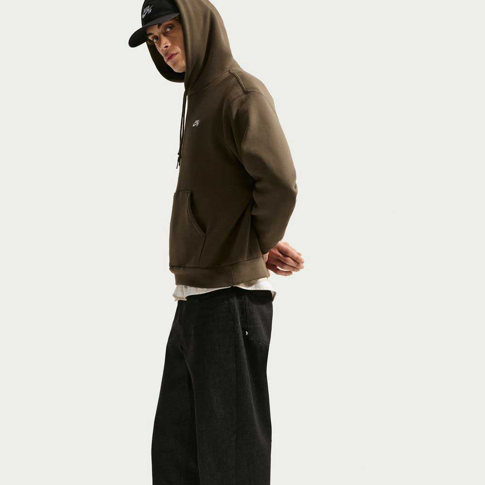A person wearing a dark green hoodie, Loose Denim Black Pant IB2992010 Nike SB, and a black Nike SB baseball cap stands sideways with hands behind their back against a plain white background.