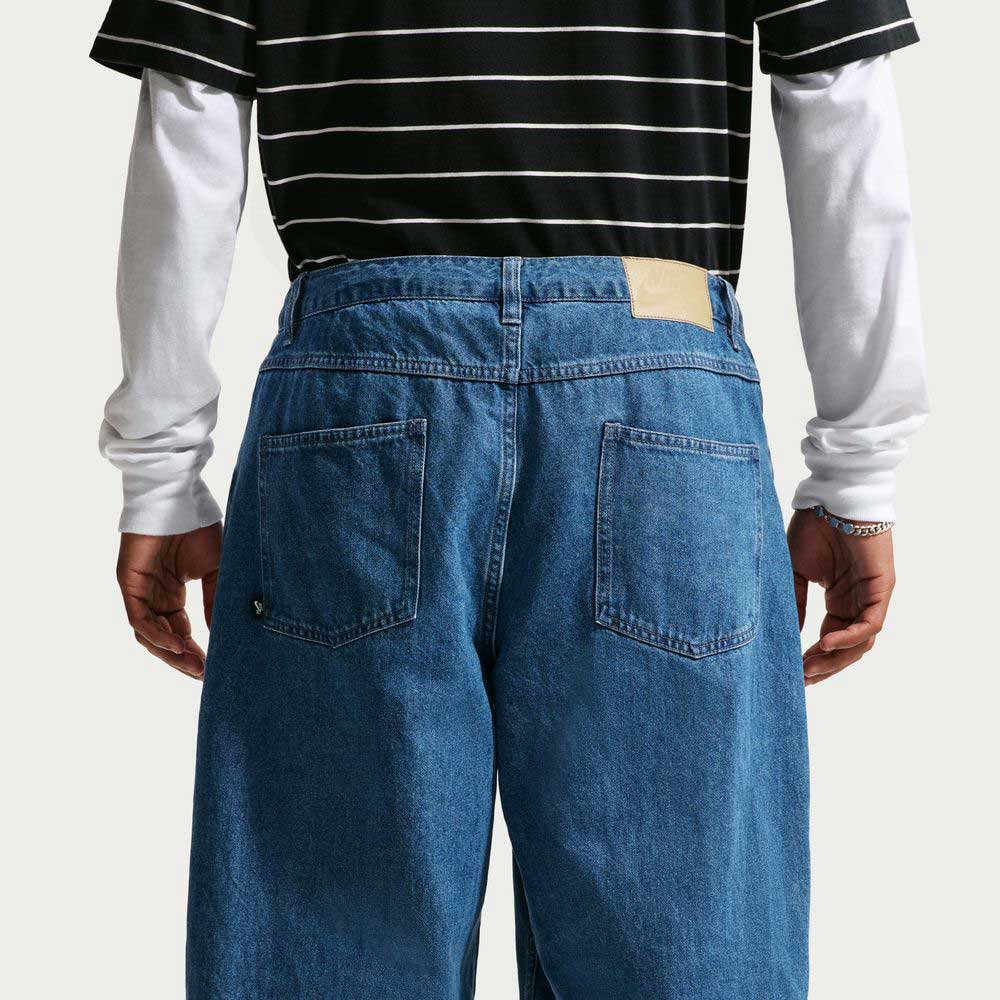 A person is seen from the back wearing Nike SB Loose Denim French Blue Pant II7363487, paired with a black-and-white striped cotton t-shirt over a white long-sleeve shirt, their hands relaxed at their sides.