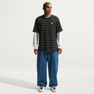 A person stands slightly turned, wearing a black and white striped 100% cotton tee over a long-sleeved white shirt, Loose Denim French Blue Pant II7363487 by Nike SB, and Nike SB white sneakers against a plain light background.