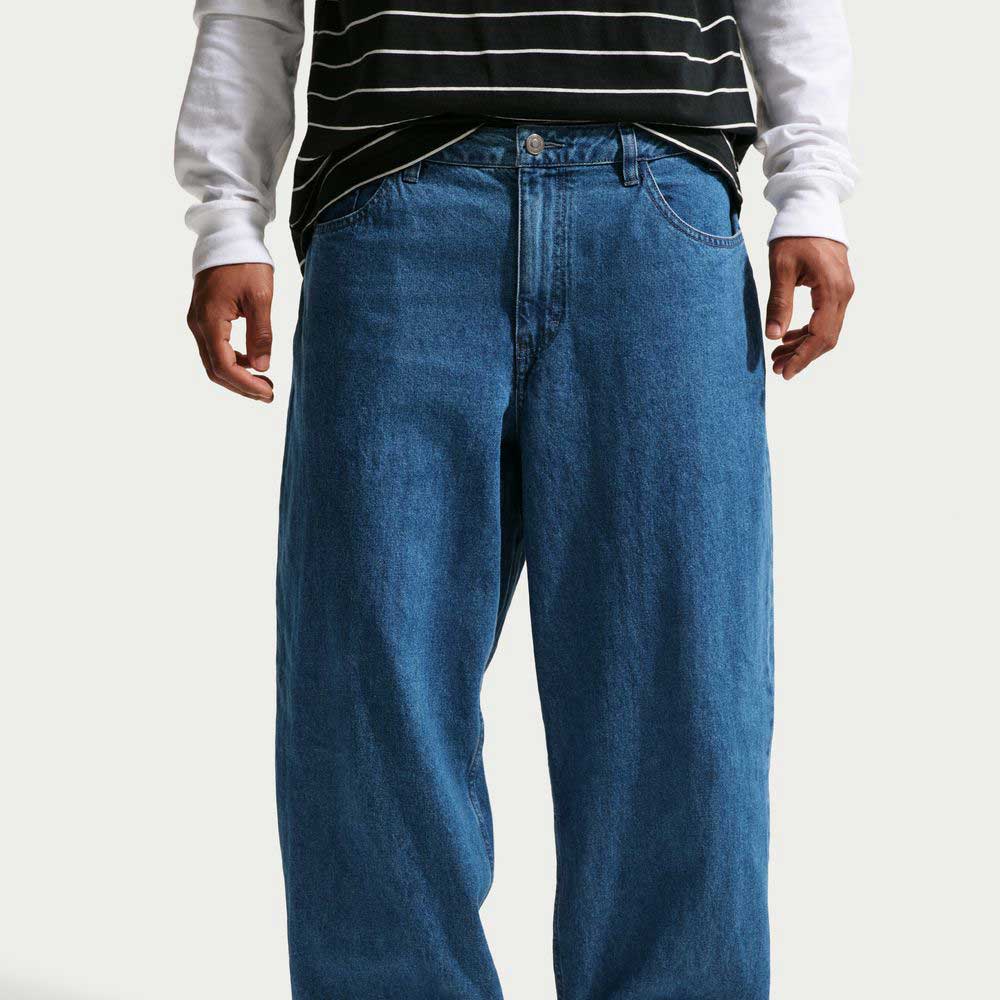 Wearing Loose Denim French Blue Pant II7363487 Nike SB, a person stands against a plain background with hands relaxed by their sides, paired with a black and white striped T-shirt and a white long-sleeve shirt.