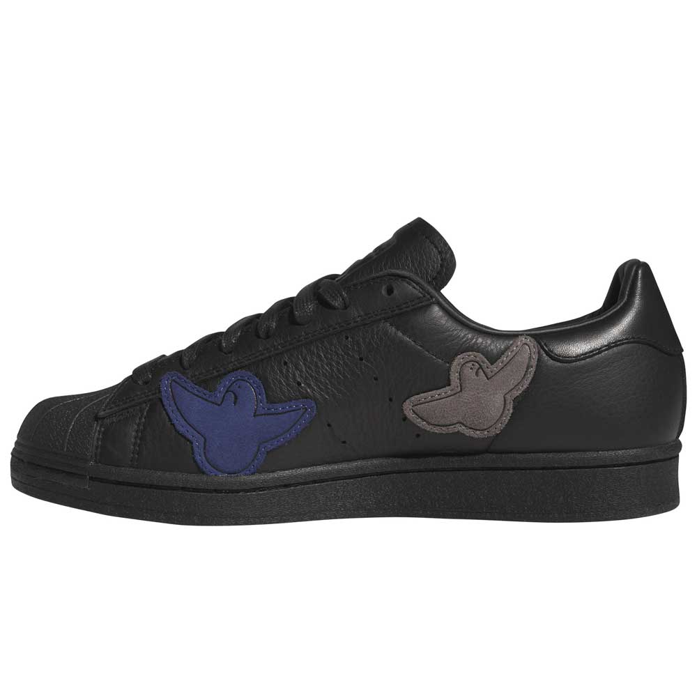 The Mark Gonzales Black Black Black LA6416 Superstar ADV x Gonz Adidas Skateboarding Leather Shoes feature black uppers with matching black laces and are accented by blue and gray bird-shaped patches on the side. Displayed in a left side profile view against a white background, these shoes offer a distinctive take on classic skateboarding style.