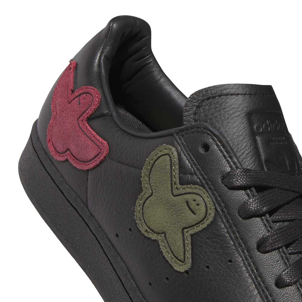 Here’s a rewritten sentence using the provided product data: A close-up of the Mark Gonzales Black Black Black LA6416 Superstar ADV x Gonz Adidas Skateboarding Leather Shoes Footwear showcases textured black leather construction, oversized suede butterfly patches in olive green and burgundy adorning the side and heel, black laces, and visible stitching—designed as premium skateboarding shoes from adidas Originals.