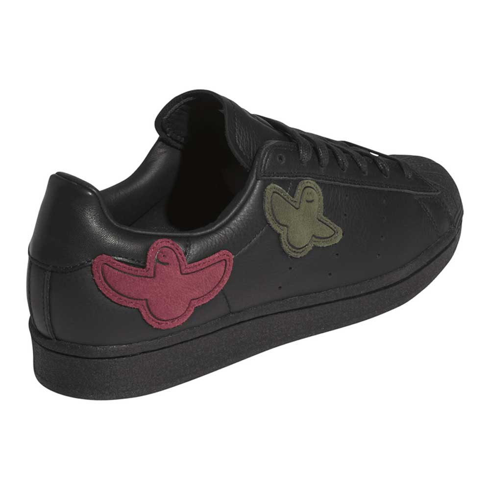 A black leather sneaker featuring red and green bird-shaped patches on the side, viewed from the back left angle, the Mark Gonzales Black Black Black LA6416 Superstar ADV x Gonz Adidas Skateboarding Leather Shoes Footwear reimagines classic adidas Originals Superstar ADV style with unique detailing inspired by skate culture.