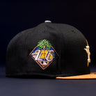 The Midsummer Night 26 Spring Bronze Houston Astros New Era Hat 5950 Fitted is a black cap with a yellow brim, featuring a colorful embroidered Astros logo with palm trees, a sunset, and "25" above the MLB logo.