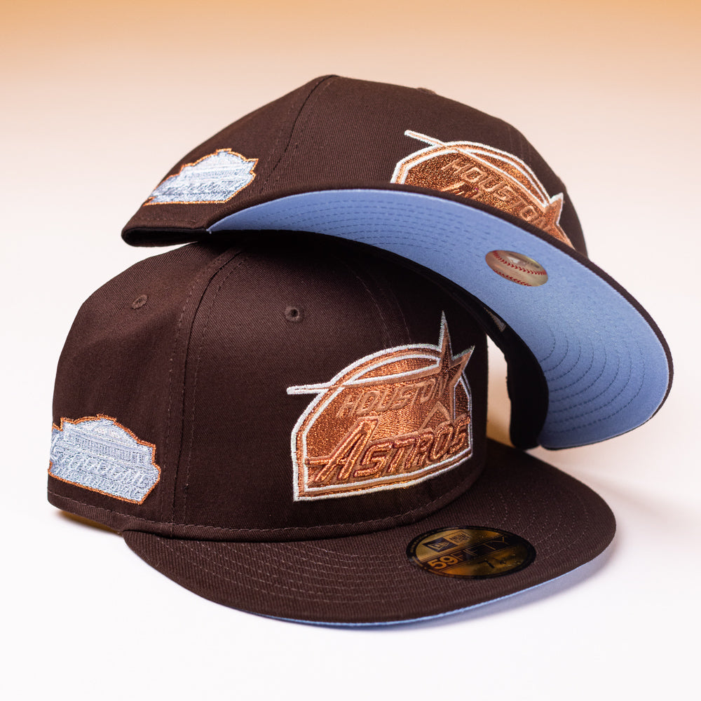Two Muddy Waters Houston Astros New Era Hat 5950 Fitted hats in brown with pale blue brims and embroidered logos are stacked; one hat is upright while the other balances on top with its brim facing upward to reveal a blue underside.