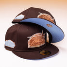 Two Muddy Waters Houston Astros New Era Hat 5950 Fitted hats in brown with pale blue brims and embroidered logos are stacked; one hat is upright while the other balances on top with its brim facing upward to reveal a blue underside.