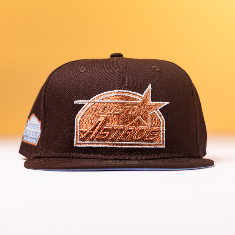 The Muddy Waters Houston Astros New Era Hat 5950 Fitted, in brown, is displayed against a yellow background and features a large embroidered gold and white team logo on the front, showcasing both style and team pride.