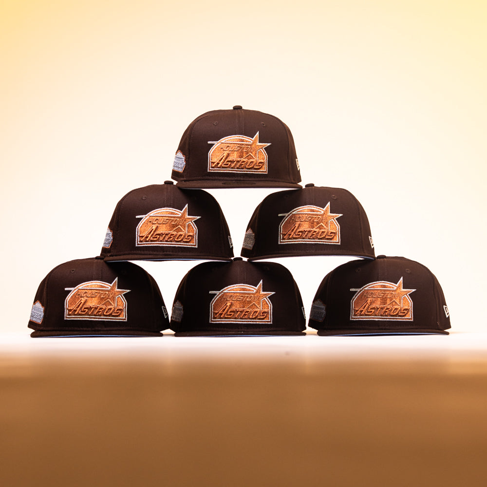 Six Muddy Waters Houston Astros New Era 5950 Fitted hats are stacked in a pyramid shape on a smooth surface, set against a warm yellow gradient background.