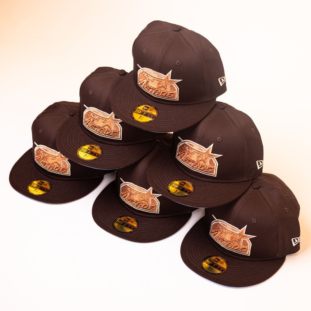 Six Muddy Waters Houston Astros New Era 5950 Fitted hats, featuring black fabric with brown embroidered Houston Astros logos and gold stickers, are stacked in a pyramid shape on a white background.