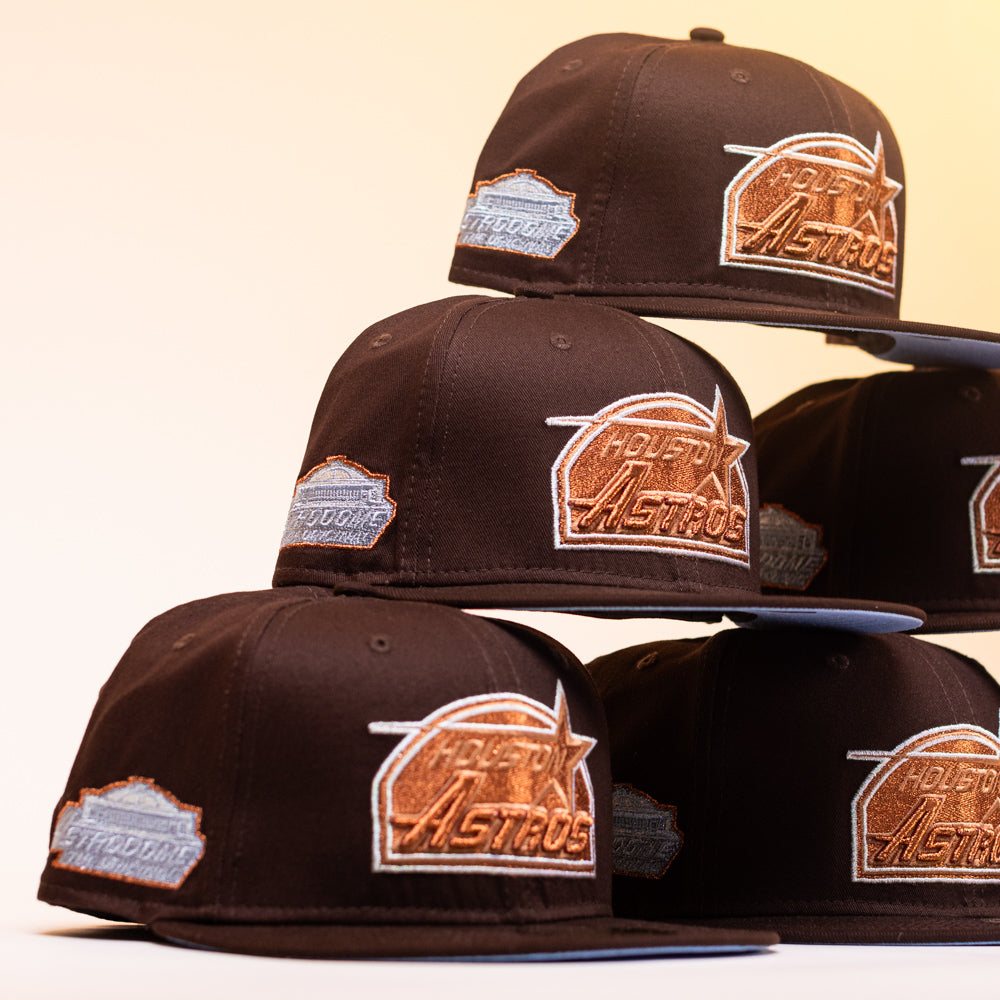 Six black Muddy Waters Houston Astros New Era Hat 5950 Fitted baseball caps with copper logos and silver patches are stacked in two uneven columns against a plain, soft beige background.