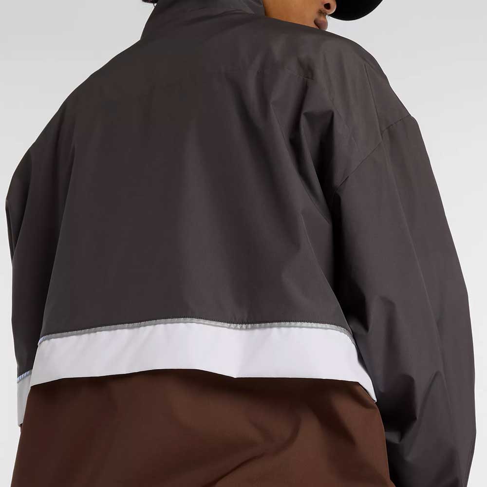 A person wearing the New Balance Numeric Jacket MJ61W4XCABR in dark gray and brown ripstop, featuring a white horizontal stripe across the back, shown from behind and slightly turned to the side.
