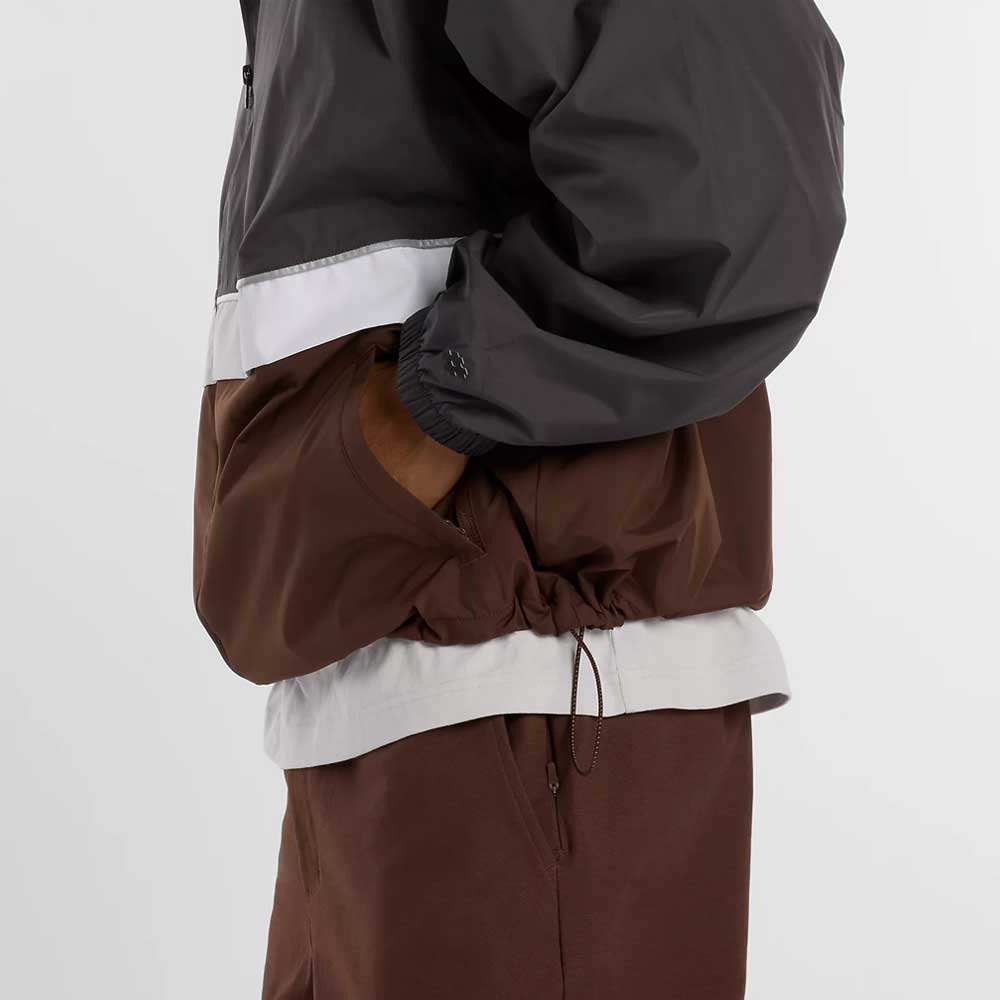A person wearing the New Balance Numeric Jacket MJ61W4XCABR in gray and brown ripstop fabric with matching brown pants, shown from the side with one hand in the jacket pocket.