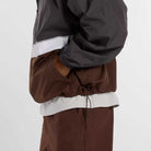 A person wearing the New Balance Numeric Jacket MJ61W4XCABR in gray and brown ripstop fabric with matching brown pants, shown from the side with one hand in the jacket pocket.