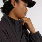 A person in profile wears a black cap and the New Balance Numeric Jacket MJ61W4XCABR in ripstop fabric, with "numeric" near the zipper. They rest their hand on their chin, showcasing gold rings and an earring.