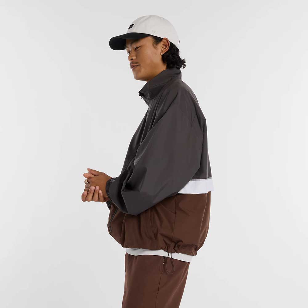 A person in a gray and brown New Balance Numeric Jacket MJ61W4XCABR made from durable ripstop fabric, brown pants, and a white baseball cap stands sideways against a plain white background.