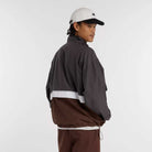 A man wearing a white baseball cap, brown pants, and the New Balance Numeric Jacket MJ61W4XCABR with NB DRY technology and a white stripe stands against a plain white background, looking over his shoulder.