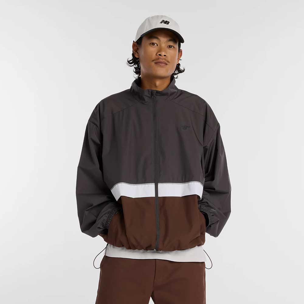 A person wearing a light gray baseball cap and a New Balance Numeric Jacket MJ61W4XCABR in dark gray, white, and brown ripstop colorblock stands with hands in pockets against a plain light background.