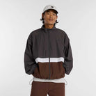 A person wearing a light gray baseball cap and a New Balance Numeric Jacket MJ61W4XCABR in dark gray, white, and brown ripstop colorblock stands with hands in pockets against a plain light background.