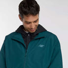 A man in a dark teal New Balance Numeric Jacket MJ61W4XCAAV with NB DRY technology and a black hoodie looks down and smiles against a plain light background.