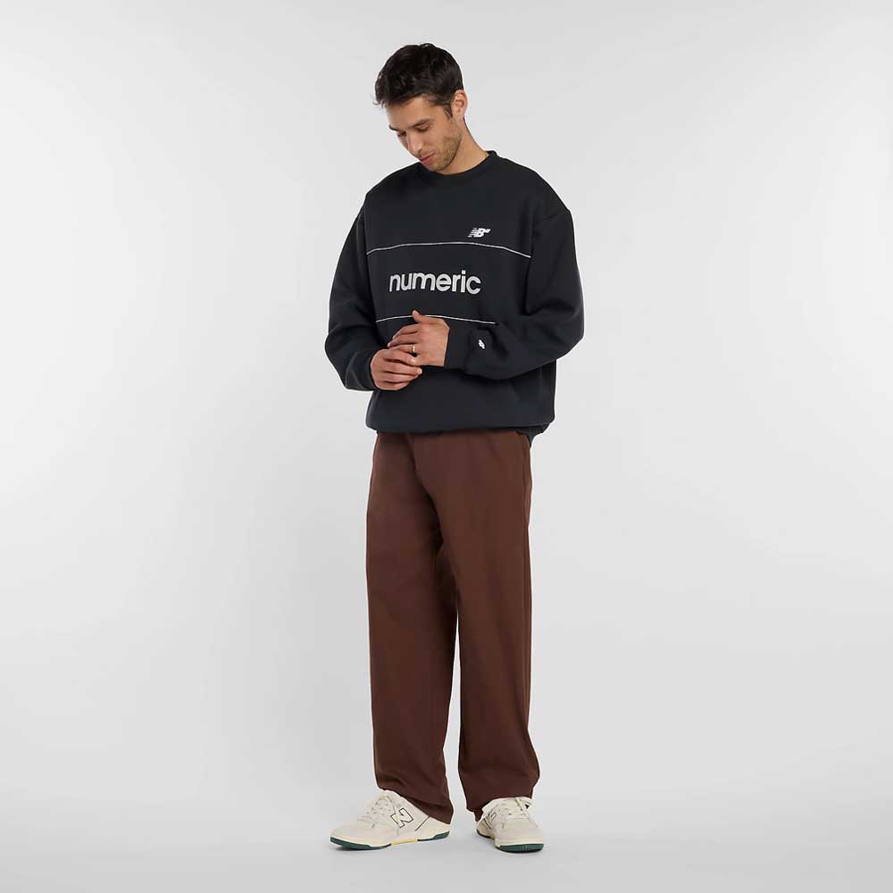 A man stands facing slightly downward, wearing a black numeric sweatshirt, New Balance Numeric Pant Pumperknickel MP51953ABJ, and white sneakers against a plain white background.