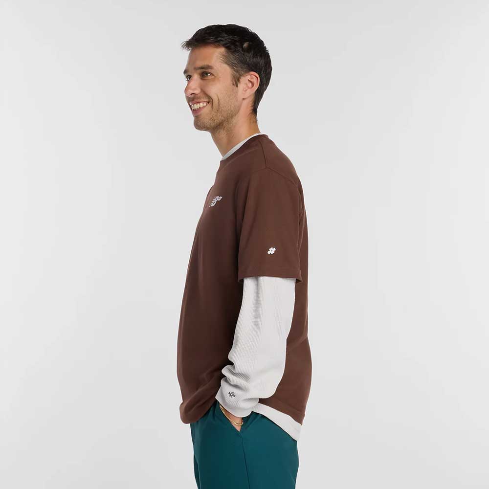 A man stands in profile, smiling with hands in his pockets. He wears the New Balance Numeric Small Logo Tee Pumpernickel MT53706ABJ over a white long-sleeved shirt and teal pants, paired with New Balance Numeric sneakers against a plain white background.