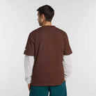 A man faces away from the camera, wearing the New Balance Numeric Small Logo Tee Pumperknickel MT53706ABJ over a white long-sleeve shirt and teal pants with Numeric sneakers, set against a plain light gray background.