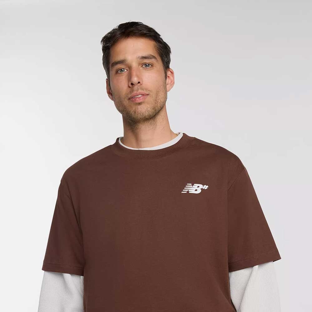 A man with short brown hair wears a relaxed fit New Balance Numeric Small Logo Tee Pumperknickel MT53706ABJ over a long-sleeve white shirt, standing against a plain light background.