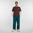A man faces forward wearing a relaxed fit brown New Balance Numeric Small Logo Tee Pumperknickel MT53706ABJ over a long-sleeve white shirt, teal pants, and white sneakers against a plain light background.