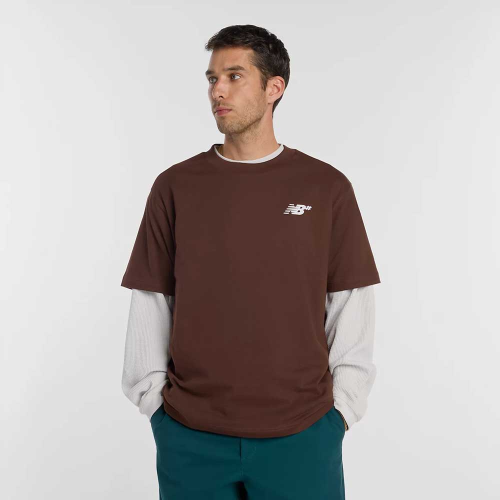 A man stands against a plain light background, wearing the New Balance Numeric Small Logo Tee Pumperknickel MT53706ABJ over a long-sleeve off-white shirt and teal pants. He looks slightly to his right with his hands in his pockets.