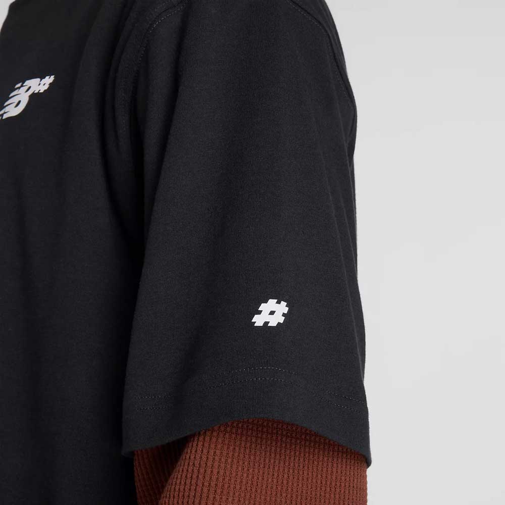 A person wears the New Balance Numeric Small Logo Black Tee MT53706BK, a relaxed fit black cotton crewneck with a small white logo on the sleeve, layered over a textured brown long-sleeve shirt against a plain background. Only part of the tee and arm is visible.