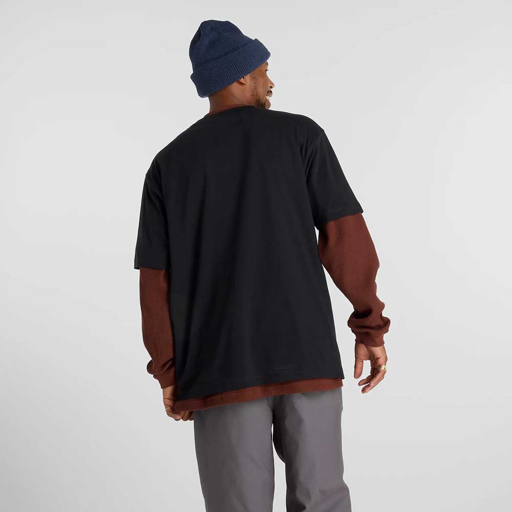 A man, facing away, wears a navy beanie, gray pants, New Balance Numeric sneakers, and the New Balance Numeric Small Logo Black Tee MT53706BK over a reddish-brown long-sleeve shirt against a plain light background.