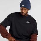 A man in a New Balance Numeric Small Logo Black Tee MT53706BK over a long-sleeve brown shirt and a blue New Balance beanie looks down with a neutral expression against a plain background.