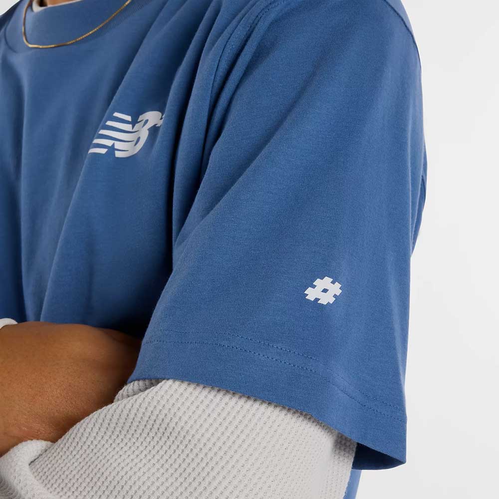 A person with crossed arms wears the New Balance Numeric Small Logo Magic Blue Tee MT53706AA4 layered over a white textured long sleeve, showing off its relaxed fit, white logo on the chest, and small hashtag motif on the sleeve.