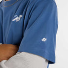 A person with crossed arms wears the New Balance Numeric Small Logo Magic Blue Tee MT53706AA4 layered over a white textured long sleeve, showing off its relaxed fit, white logo on the chest, and small hashtag motif on the sleeve.