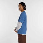 A person with long dark hair stands in profile against a white background, wearing the New Balance Numeric Small Logo Magic Blue Tee MT53706AA4 over a gray long-sleeve shirt and brown pants.