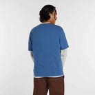 A person with dark, wavy hair faces away, wearing the New Balance Numeric Small Logo Magic Blue Tee MT53706AA4 over a long-sleeved gray shirt and brown pants, paired with New Balance Numeric sneakers against a plain white background.