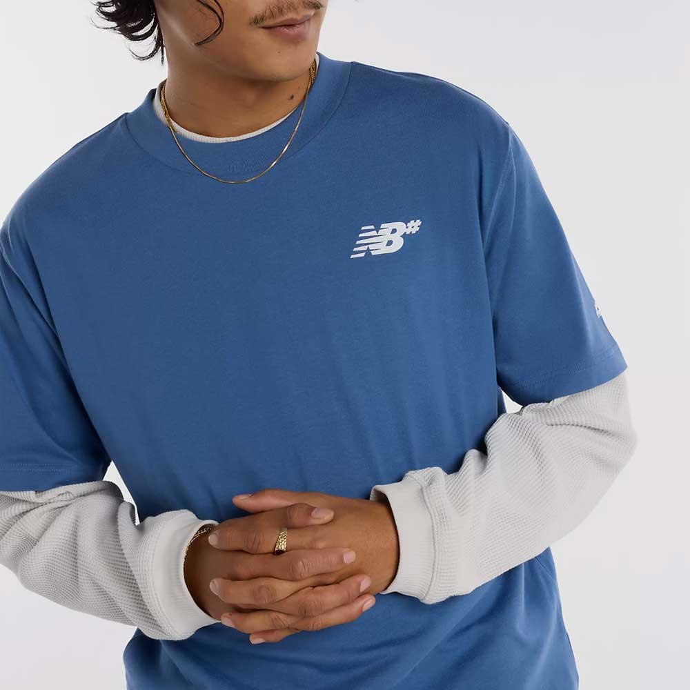 Wearing a New Balance Numeric Small Logo Magic Blue Tee (MT53706AA4) over a white long-sleeve, with a gold chain and ring, this person stands with hands clasped. The relaxed fit offers comfort; their face is partially out of frame.