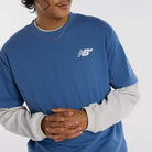 Wearing a New Balance Numeric Small Logo Magic Blue Tee (MT53706AA4) over a white long-sleeve, with a gold chain and ring, this person stands with hands clasped. The relaxed fit offers comfort; their face is partially out of frame.