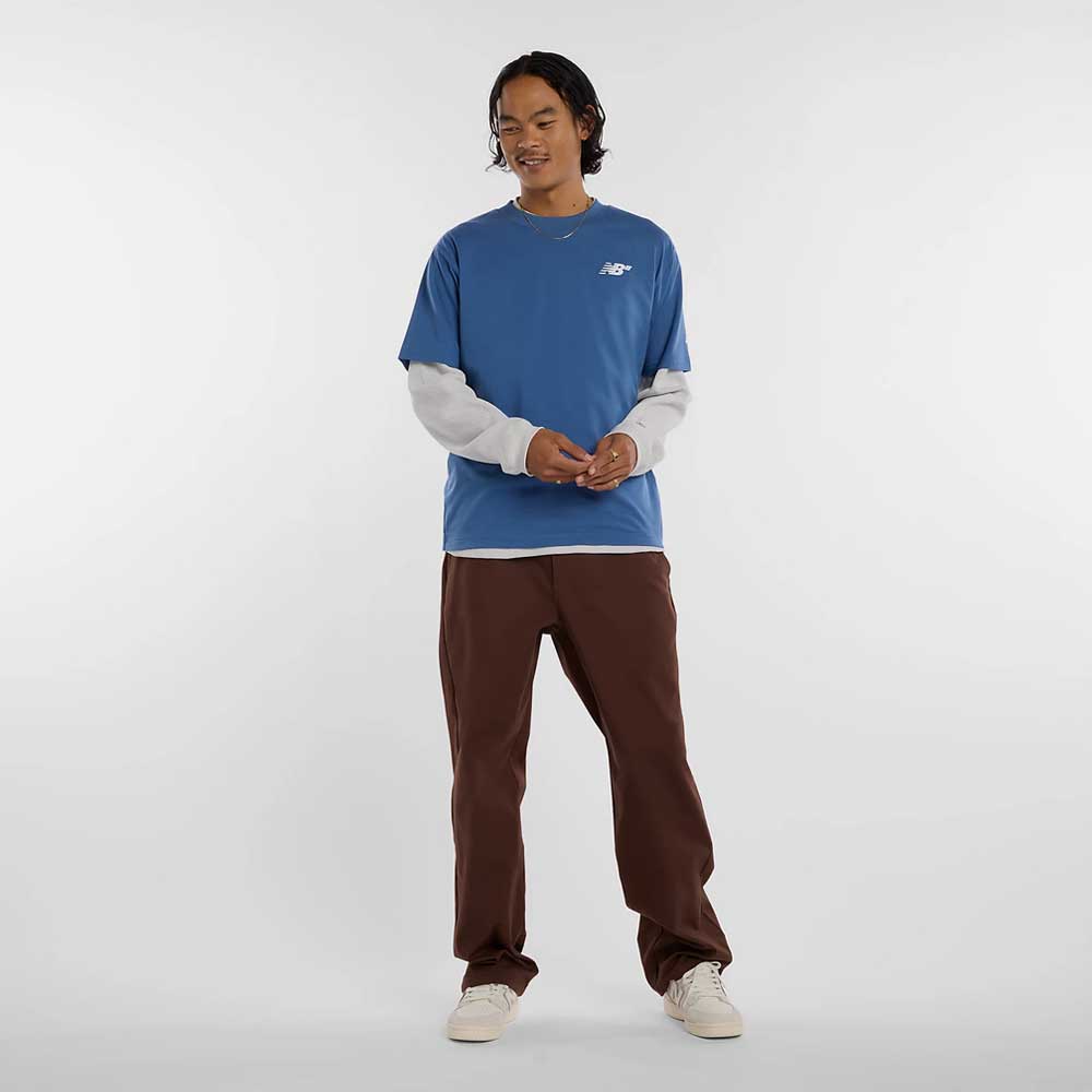 A person stands against a plain background, wearing the New Balance Numeric Small Logo Magic Blue Tee MT53706AA4 over a white long-sleeve shirt, paired with brown pants and white sneakers, looking slightly to the side and smiling.