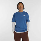 A person with wavy dark hair wears the New Balance Numeric Small Logo Magic Blue Tee MT53706AA4 over a long-sleeved gray shirt and brown pants, standing with hands in pockets against a plain white background.
