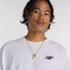 A person with wavy dark hair and a thin mustache wears a relaxed fit New Balance Numeric Small Logo White Tee MT53706WT and two gold necklaces, one with a round pendant, against a plain light background.